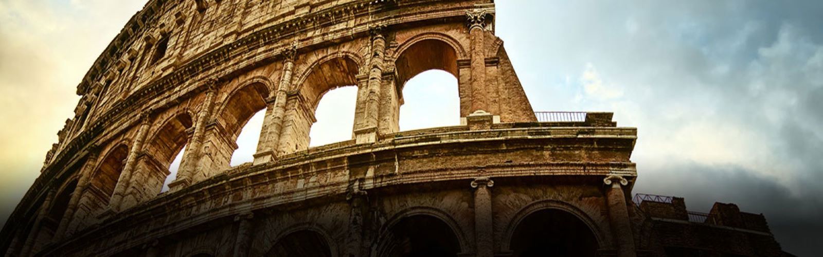 The sunrise coming over the Coliseum, a stone building with many layers and arches. 